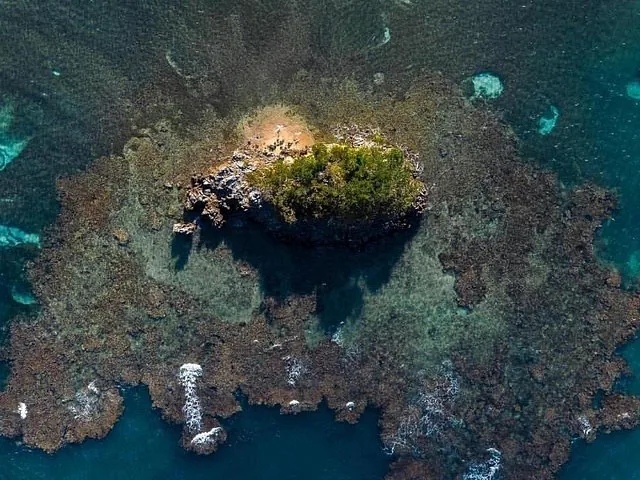 Luftaufnahme einer kleinen, felsigen Insel, umgeben von einem flachen Korallenriff.