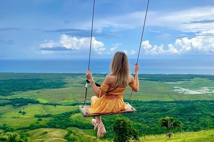 A woman in a yellow dress sits on a swing overlooking a vast green landscape.