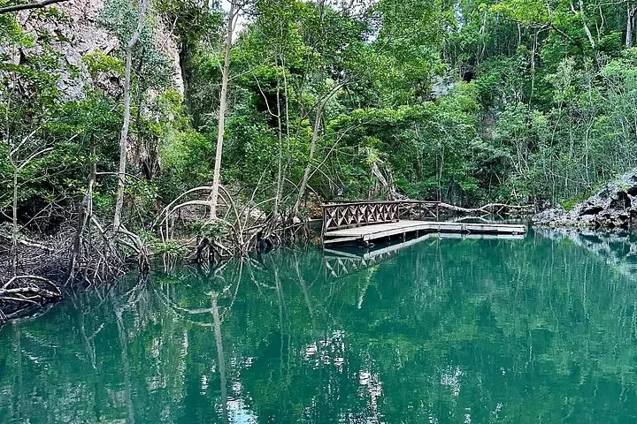 Ein hölzerner Steg erstreckt sich über klares türkisfarbenes Wasser, umgeben von Bäumen und Felsformationen.